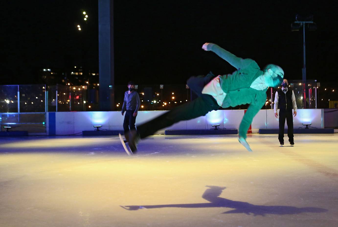 A man in a green cardigan shoots off the ice, his body is slanted, his arms and legs extended in relaxed Vs. Two other men stand behind him motionless